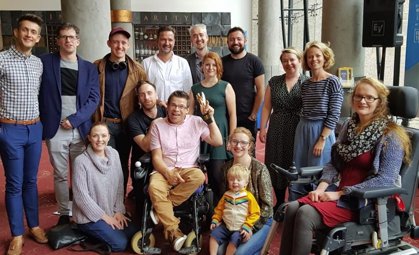 Various members of the cast and crew for My Left / Right Foot clump together in the foyer of Festival Theatre, Robert holds the Herald Angel ward up into the air. Everyone is smiling.