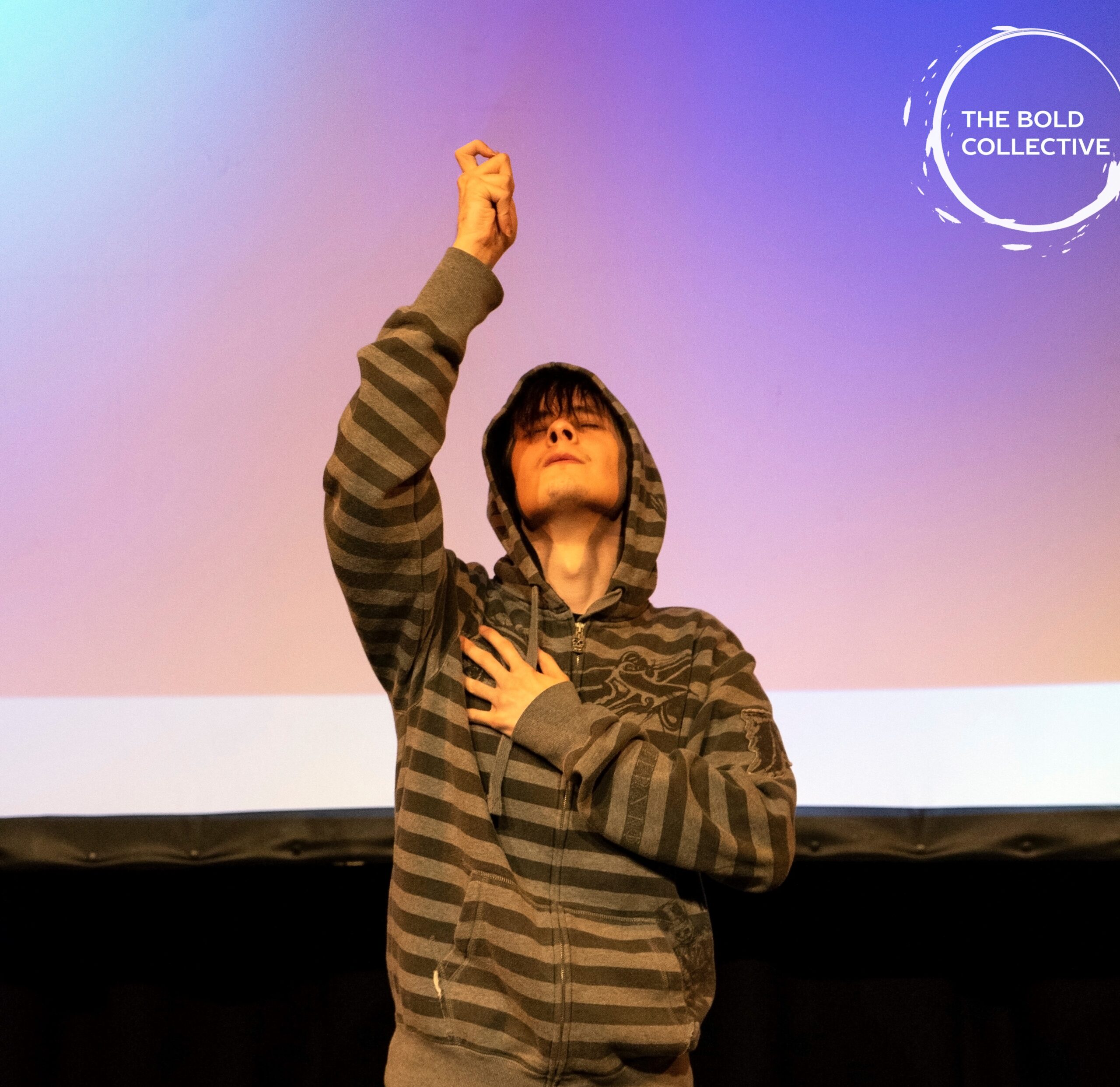 A performer- his hood is up, his eyes are closed. One hand is on his heart, his other hand reaches upwards- hopeful.