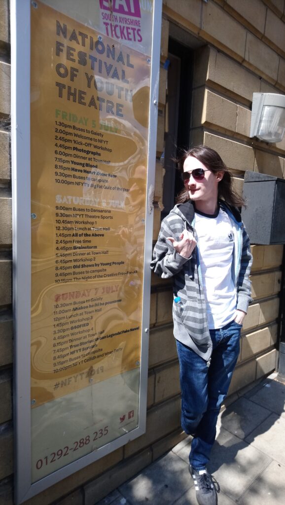 Andrew stands beside a poster which has the full listings for National Festival of Youth Theatre 2019