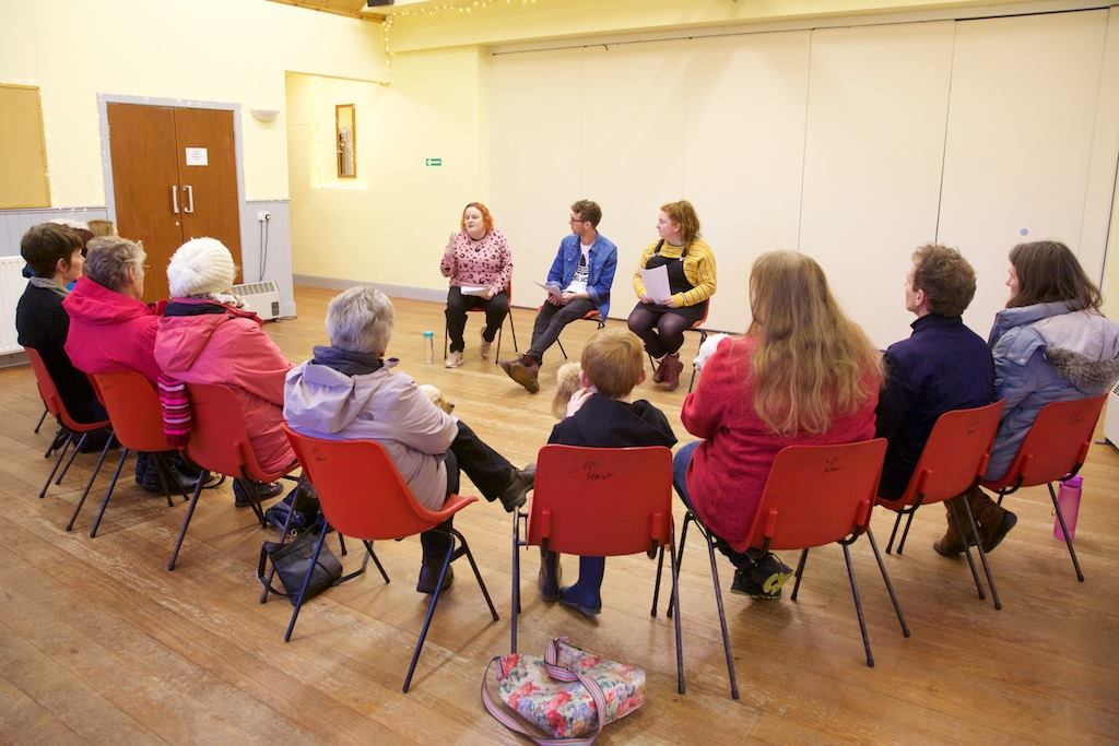 A shot from behind a large group of 13 people. They appear to be a range of ages; from childhood to old age. They are gathered in red plastic chairs in a semi circle all pointed towards three young people at the front who appear to be talking.