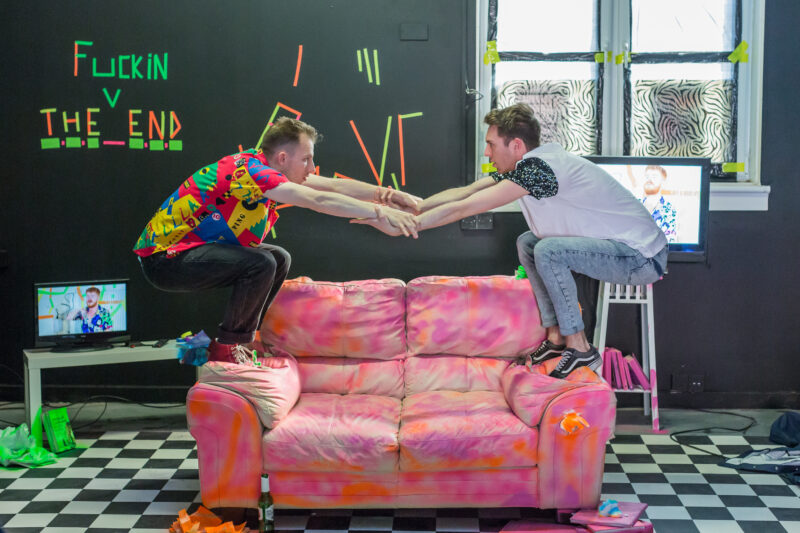 A room with black walls and a black and white checkered floor, there are zebra print blinds over the windows and stretches of brightly coloured tape everywhere. On the left hand side of the wall the phrase 'The fucking end' is spelt out in tape. In the centre there is a blotchy pink sofa. Two young men, both in: sneakers, skinny jeans and t shirts, stand on either arm of the sofa. They are reaching out and grabbing onto each others arms.