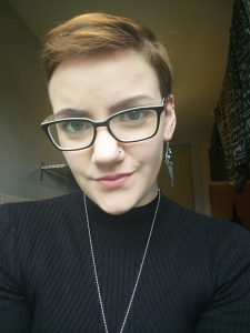 Headshot of Morna McGeoch, she is a young woman with short brown hair, glasses and a long dangly metal earing with a few other ear studs and a nose stud.