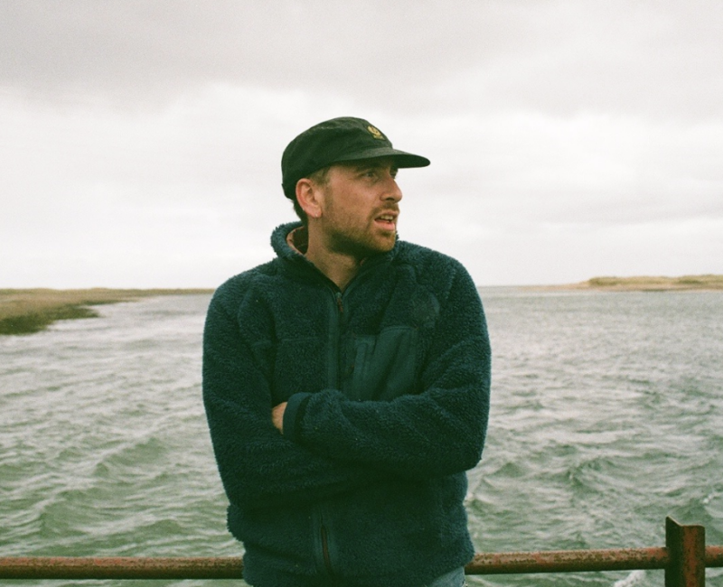 Image of the devastatingly handsome Callum. He is wearing a fluffy blue fleece and a black cap and is staring off into the distance while standing on a bridge with a murky estuary behind him.