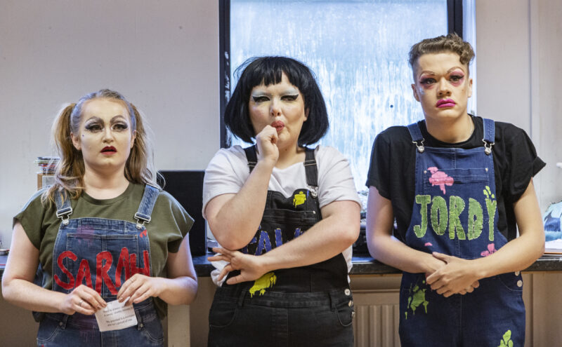 Three people in drag makeup. They are all wearing T shirts with denim dungarees, The dungarees have names on front, the girls on the left read 'Sarah' and the boys on the right read 'Jordy. The person in the middle has their arms crossed, so the name is obscured, they are putting a lollipop in their mouth.