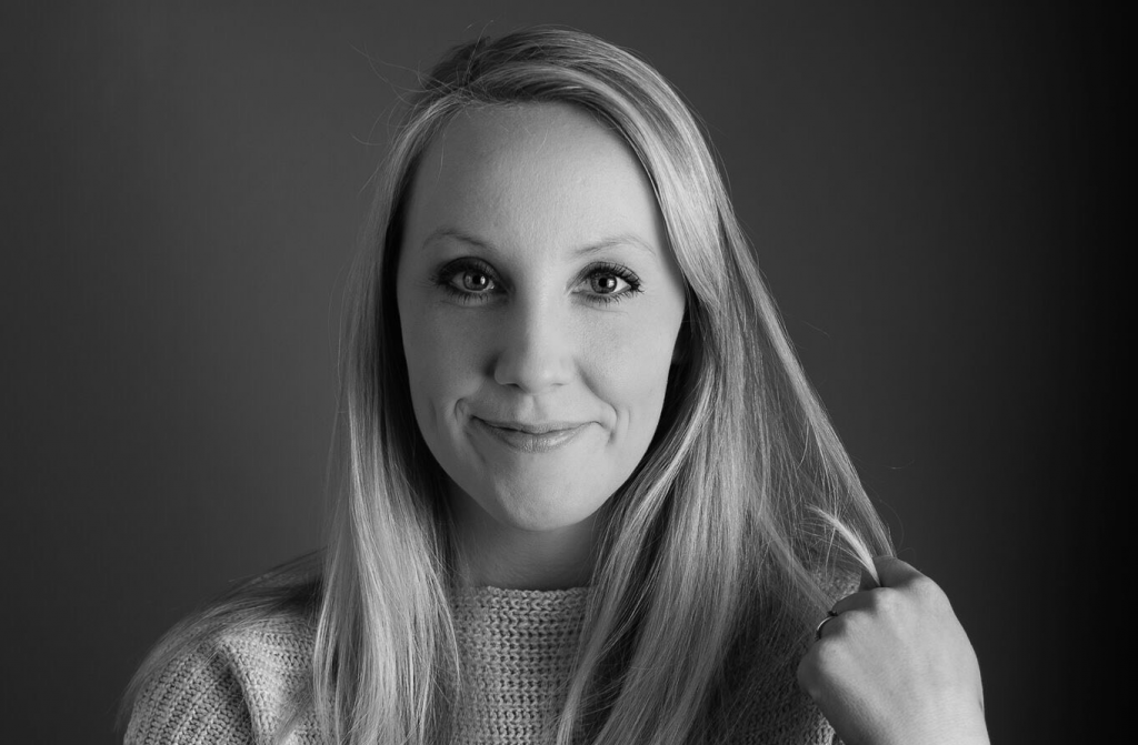 A young woman stares into the camera, she is wearing a jumper and flicks her long her with her left hand.