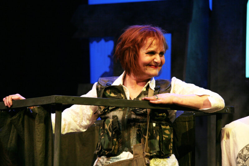 An older woman stands leaning on some railing. She has messy red brown hair and is smiling. She is wearing a white blouse with a camouflage waistcoat.