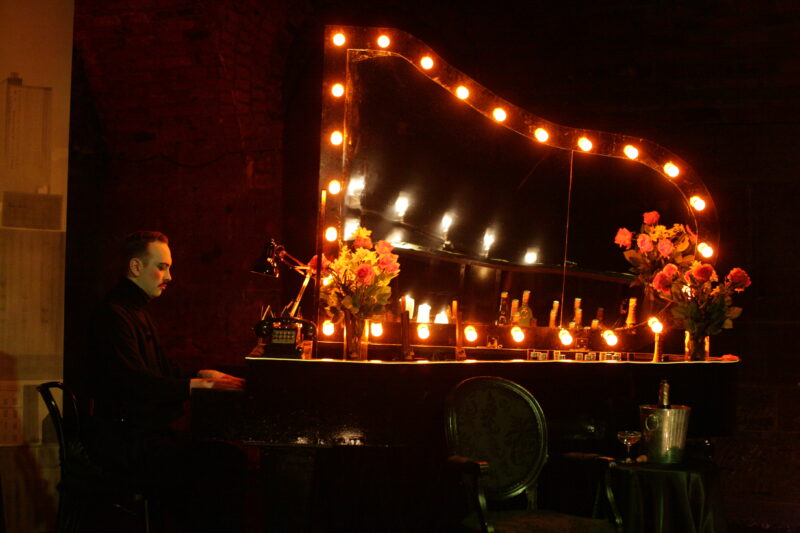 A man dressed in all black, sits playing the piano. He has slicked back dark hair and a moustache. The top of the piano is covered in lights. We also see multiple bouquets of flowers as well as a telephone on top of the piano.