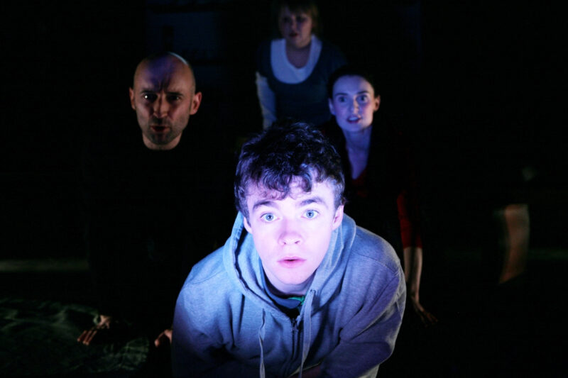 Four people gather and look at a light source off stage, they are watching it as if it is a TV. They look intrigued