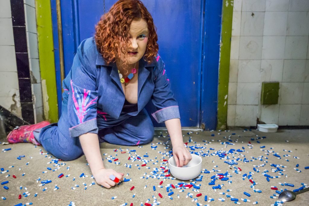 Wearing a blue boiler suit, Heather is on her hands and knees in what looks like a run down public toilet, the floor is covered in lots of pills and she is trying to collect them up into a white ceramic bowl.
