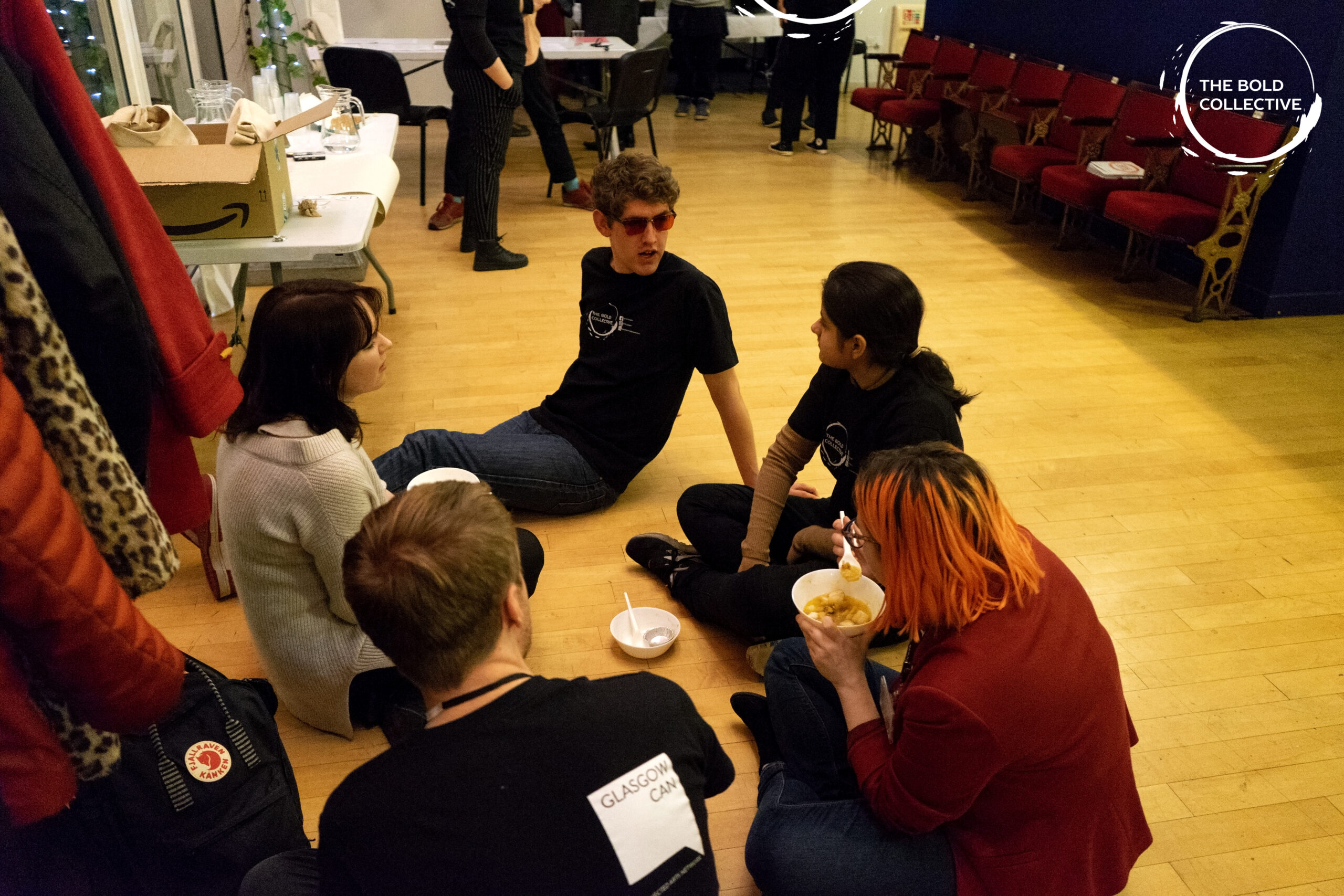 A group of five young people sit on the floor- relaxing, eating food and chatting.