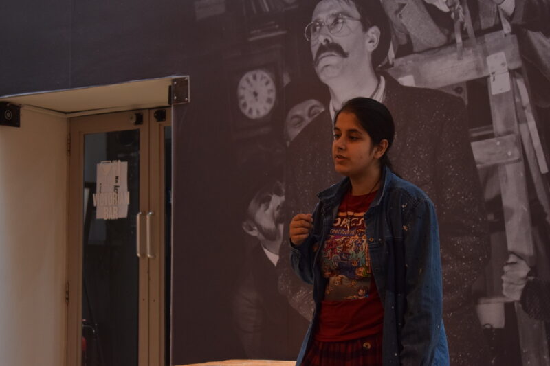 Photo of a young woman artist (Mariam) of Asian heritage standing in a room at Tron Theatre. They are alone but on the wall behind them is another picture of a larger than life figure. Mariam is in her early 20s with a dark blue shirt and marron t-shirt depicting something that looks influenced by comics.