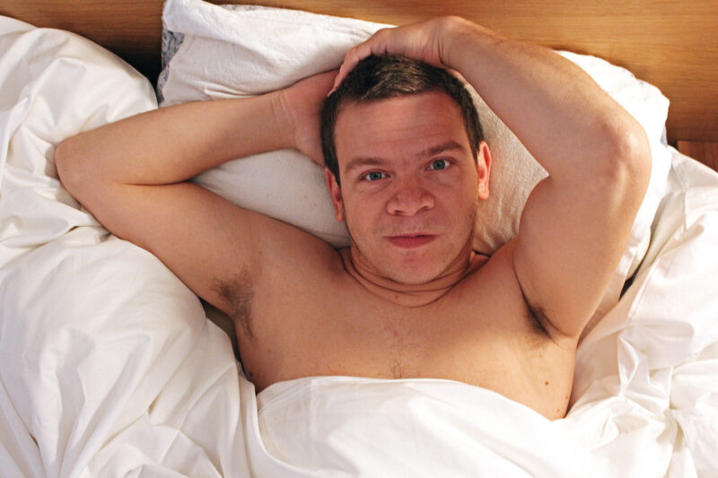 Robert, a white middle aged man, lies back in bed facing the camera. He has the white bed covers pulled up over his nipples and his hands back behind his head. He looks smoulderingly at the camera.
