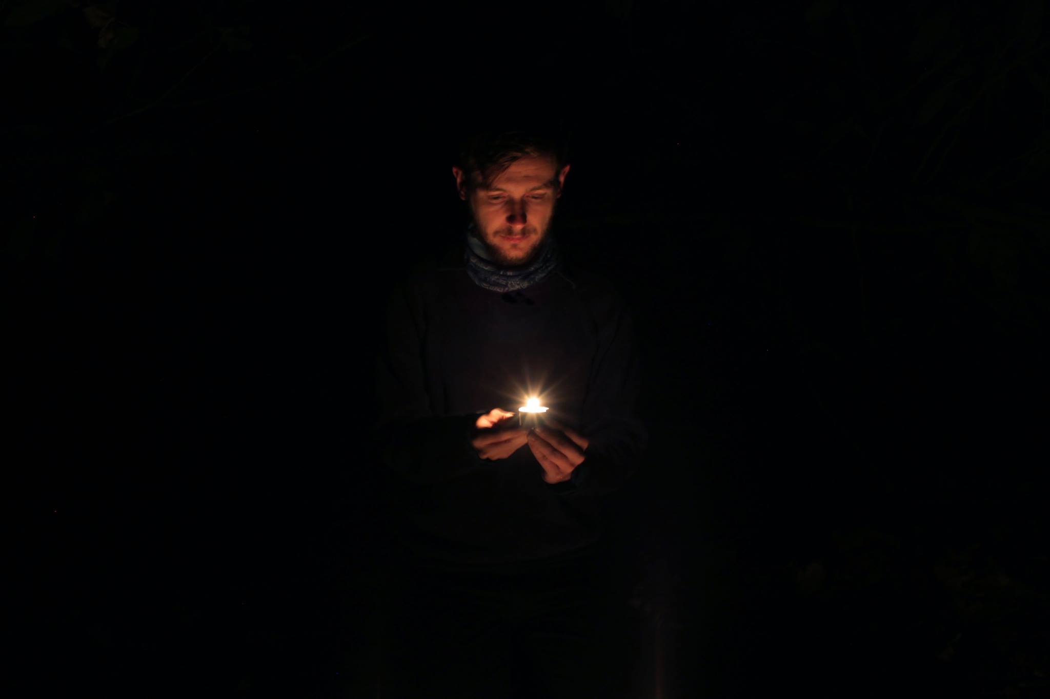 Thom Scullion looks down at a candle burning in his hands. He is a white man in his 30s. His face is bathed in an orange glow while around him is darkness.