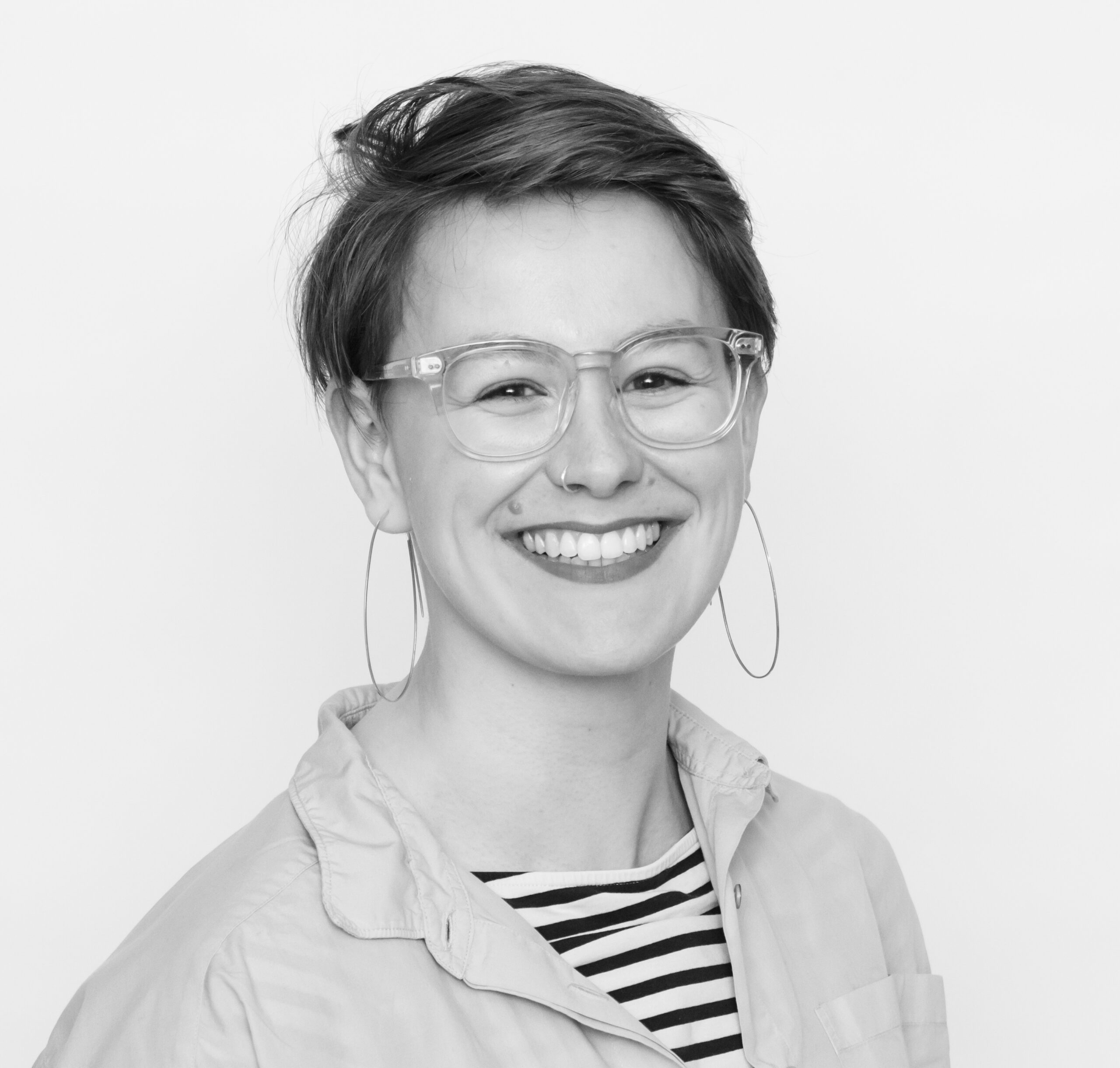 Black and white headshot of Any Watson: a white woman in her 30s with short hair. she has a shit over a stripy t-shirt, big hoop earrings, glasses and a big smile