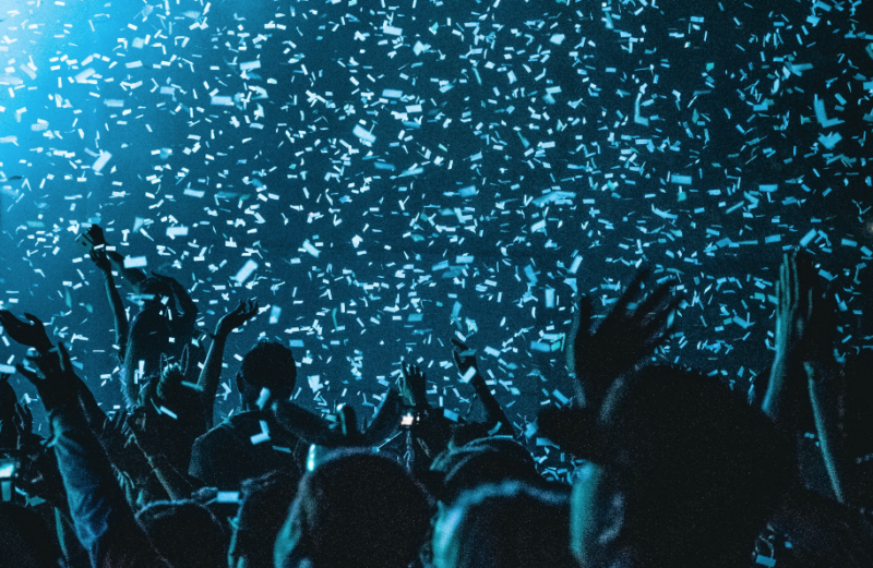 Image of a crowd from behind. all we can see are the backs of head and arms raised in the air silhouetted against a bright blue light coming from the top left. The air above the crows is filled with blue and white confetti.