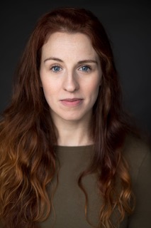 Zoe is a white woman in her mid 30s. She has long dark brown hair that hangs past her shoulders in waves. She has one eyebrow partially raised in a friendly manner.