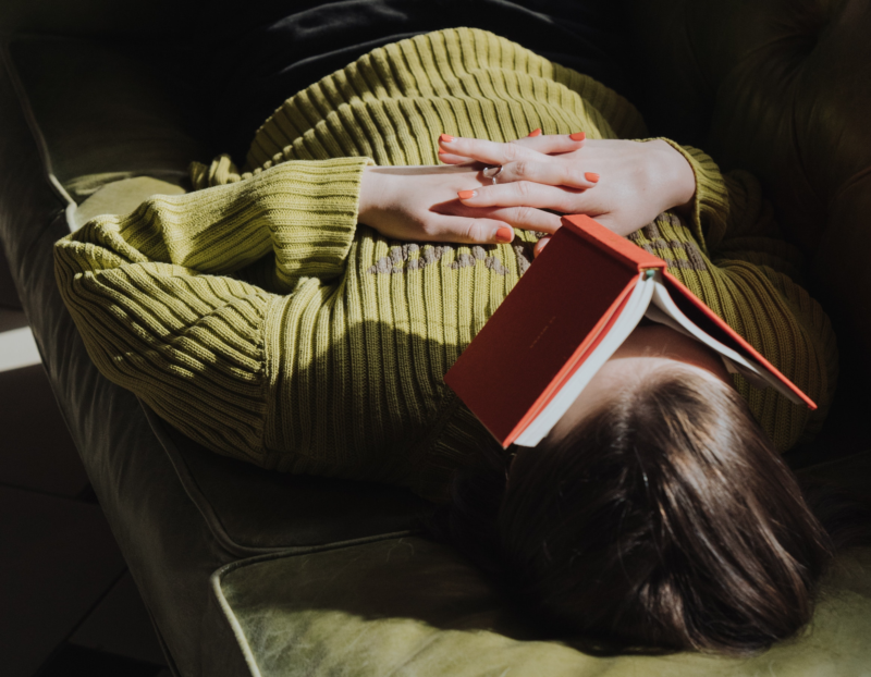 A woman lies on her back on a sofa, hands clasped across her chest. A book with a red cover is open, lying across her face.