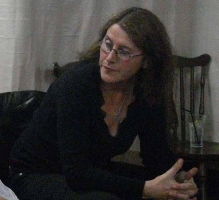 Aine is a white middle aged woman with long brown hair and glasses. In this picture she is perched on the edge of a chair her hands clasped together, while she concentrates on something to the side.