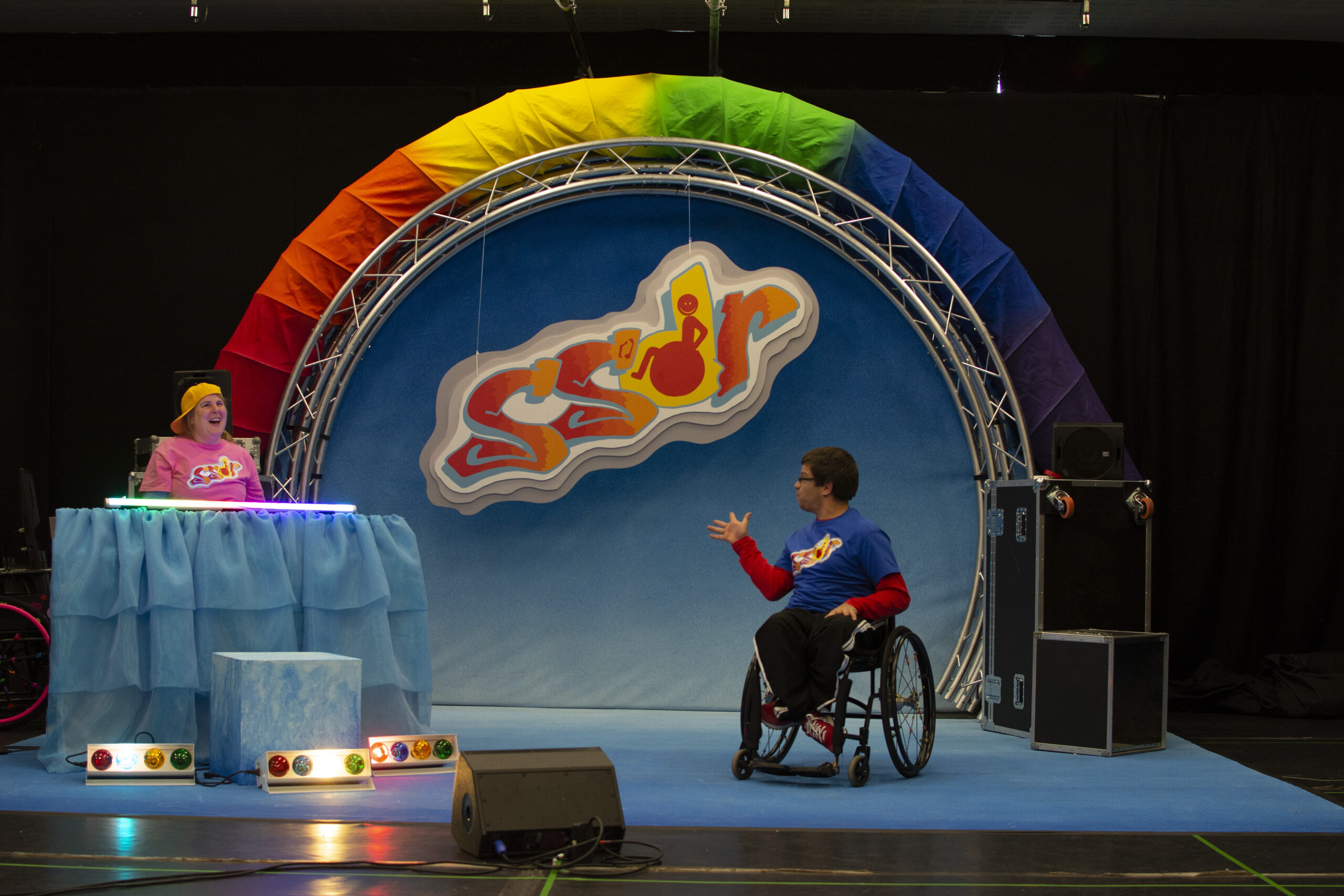 Sally stands behind the music deck with the fluorescent light on top of it. On the floor in front of the desk, there are 3 long lights with 4 different coloured bulbs in each of them: red, yellow, green and white. The white bulbs are lit up. Robert is near the front of the stage and is looking back at Sally with one of his hands opened and raised. 