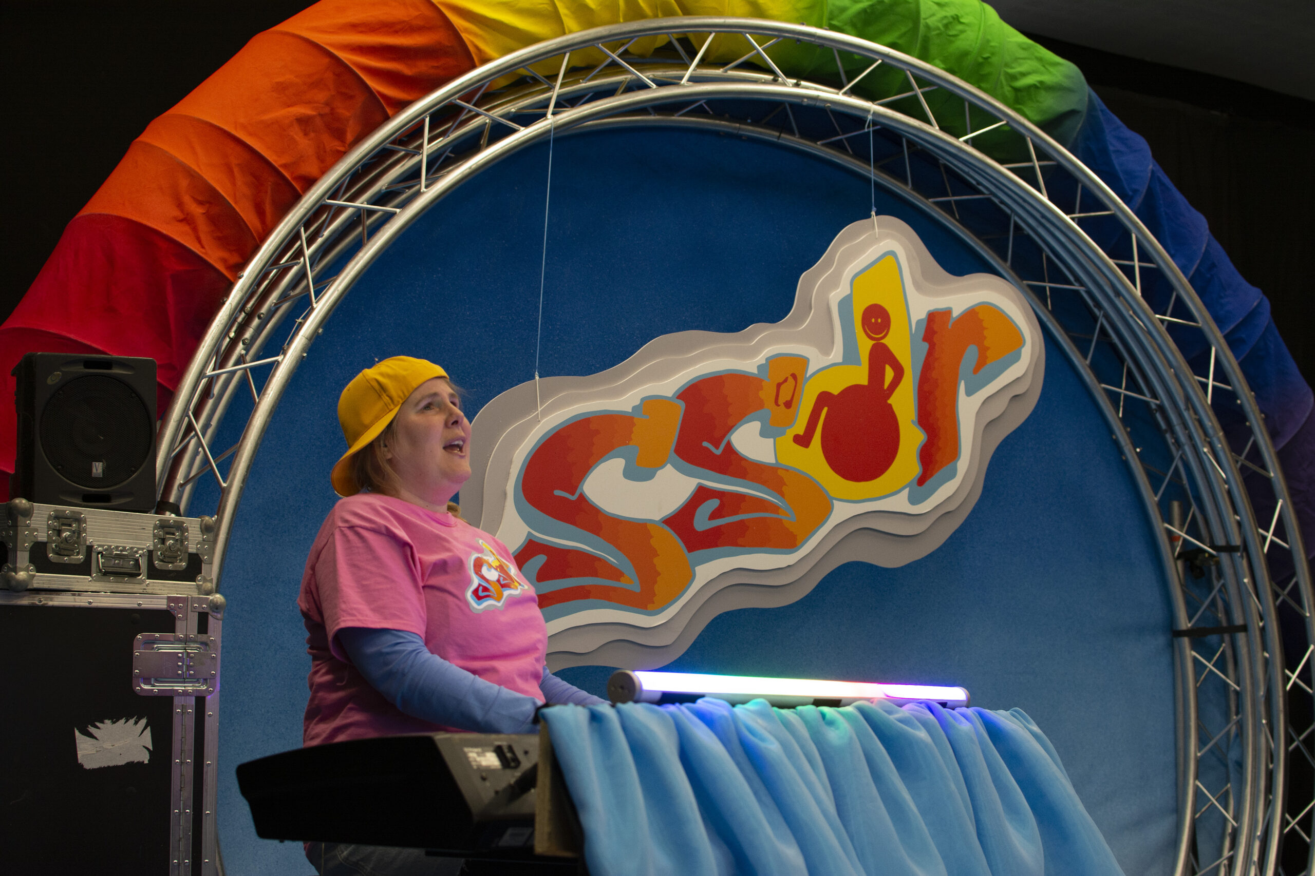 A close up of Sally behind the music deck. She appears to have a keyboard in front of her. Her mouth is open as she appears to be singing. 