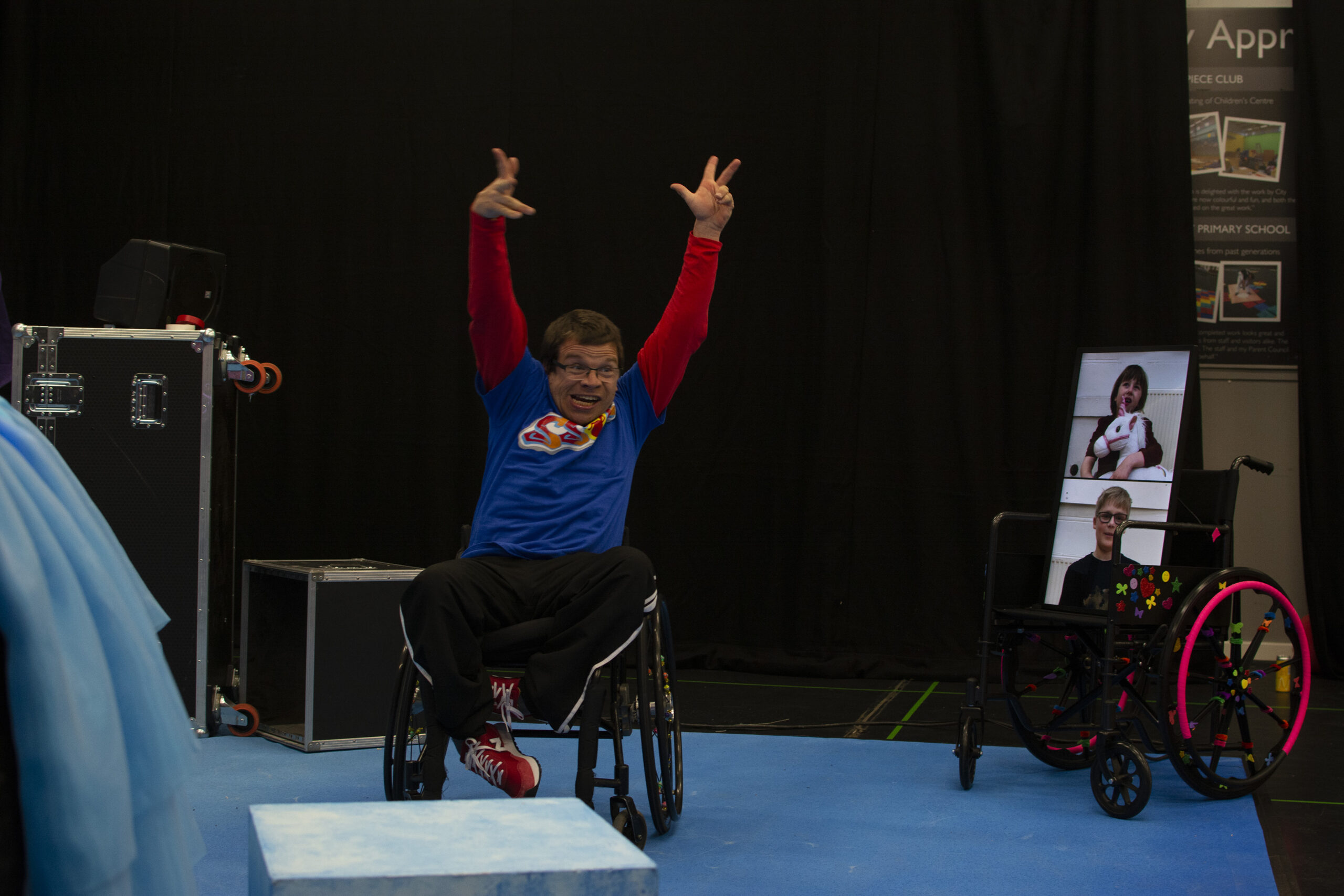 A shot of Robert near the side of the stage. He has both of his arms stretched up high in the air. To the right of him, there is the screen on a wheelchair. The wheelchair has various brightly coloured stickers all over it. The screen is split with video feeds of Ollie and Oona, at the top is Oona with a dark bob haircut holding a white toy unicorn. Beneath her is Ollie. 