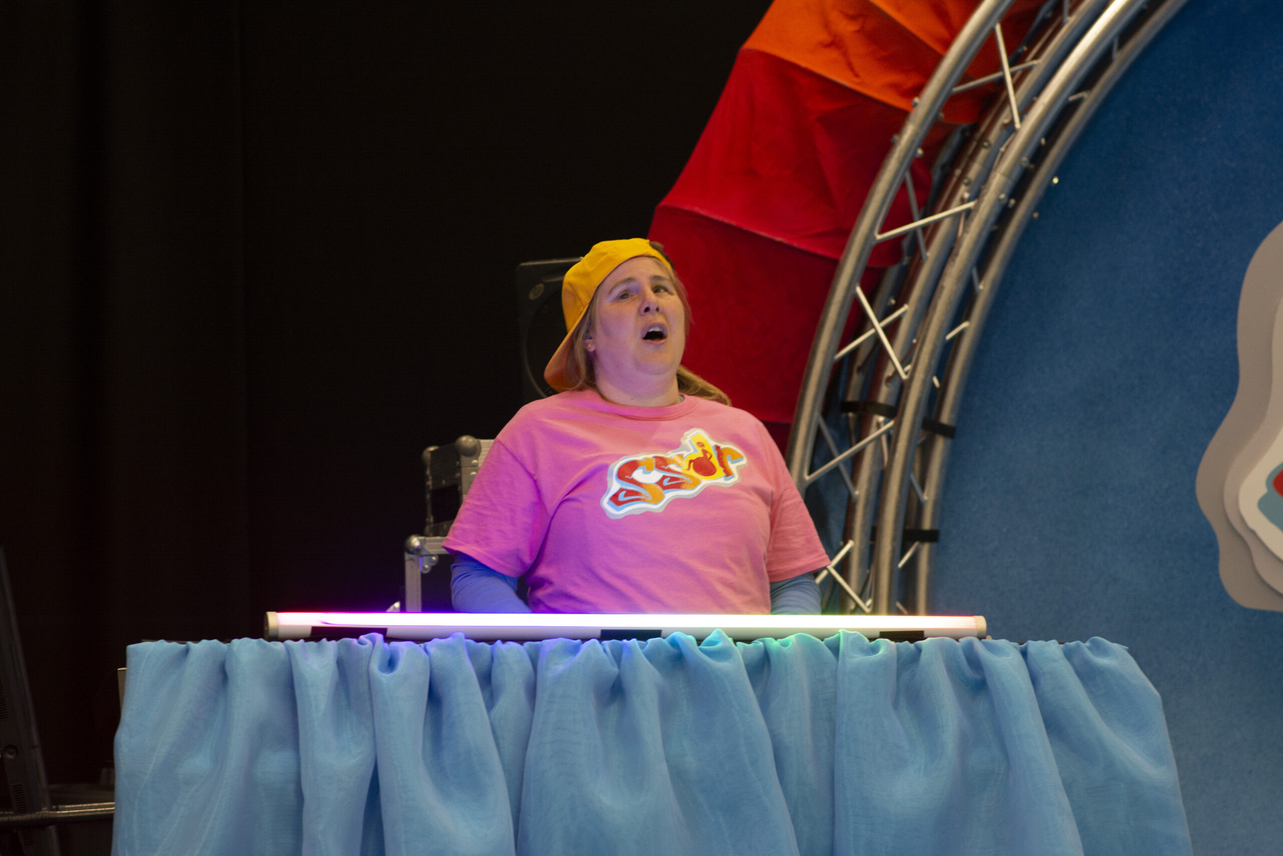 Sally stands behind the music desk. There is a long fluorescent light bulb running across the top of it that lights her face. It is giving of a lilac glow. Her mouth is open and she appears to be mid song. 