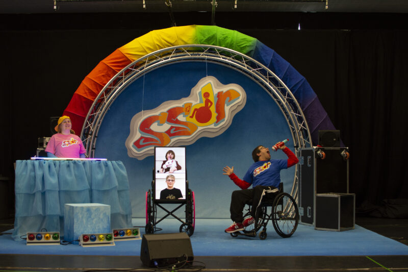 The woman is standing on the left behind the musical equipment that is covered in a blue sheet. She is smiling. On the right, the man in the wheelchair is holding a microphone with his mouth wide open and his head thrown back. In the middle of them, there is a screen on wheels with two people on it against a white background. One of these people appears to be holding a white toy pony.