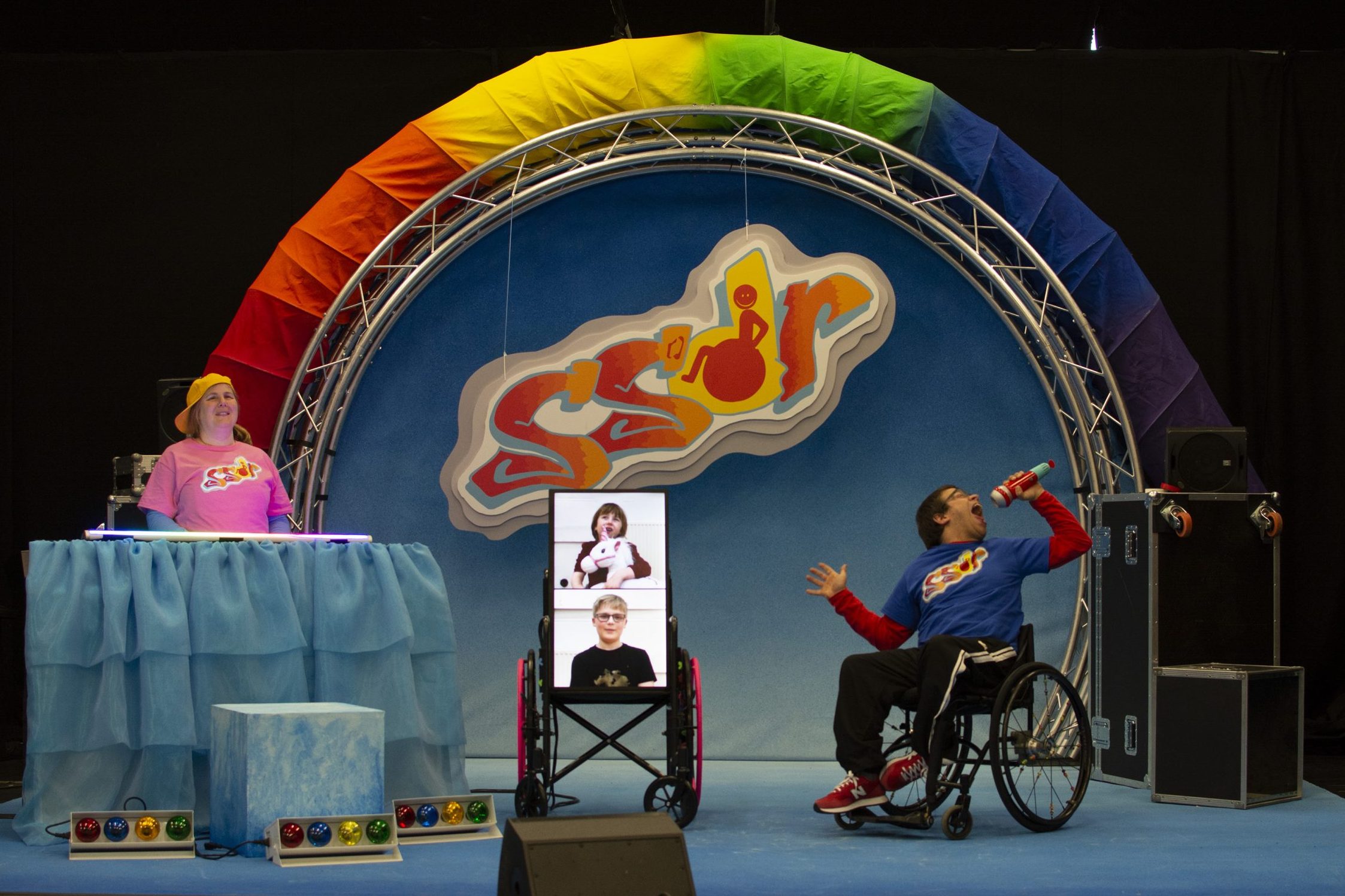 The woman is standing on the left behind the musical equipment that is covered in a blue sheet. She is smiling. On the right, the man in the wheelchair is holding a microphone with his mouth wide open and his head thrown back. In the middle of them, there is a screen on wheels with two people on it against a white background. One of these people appears to be holding a white toy pony.