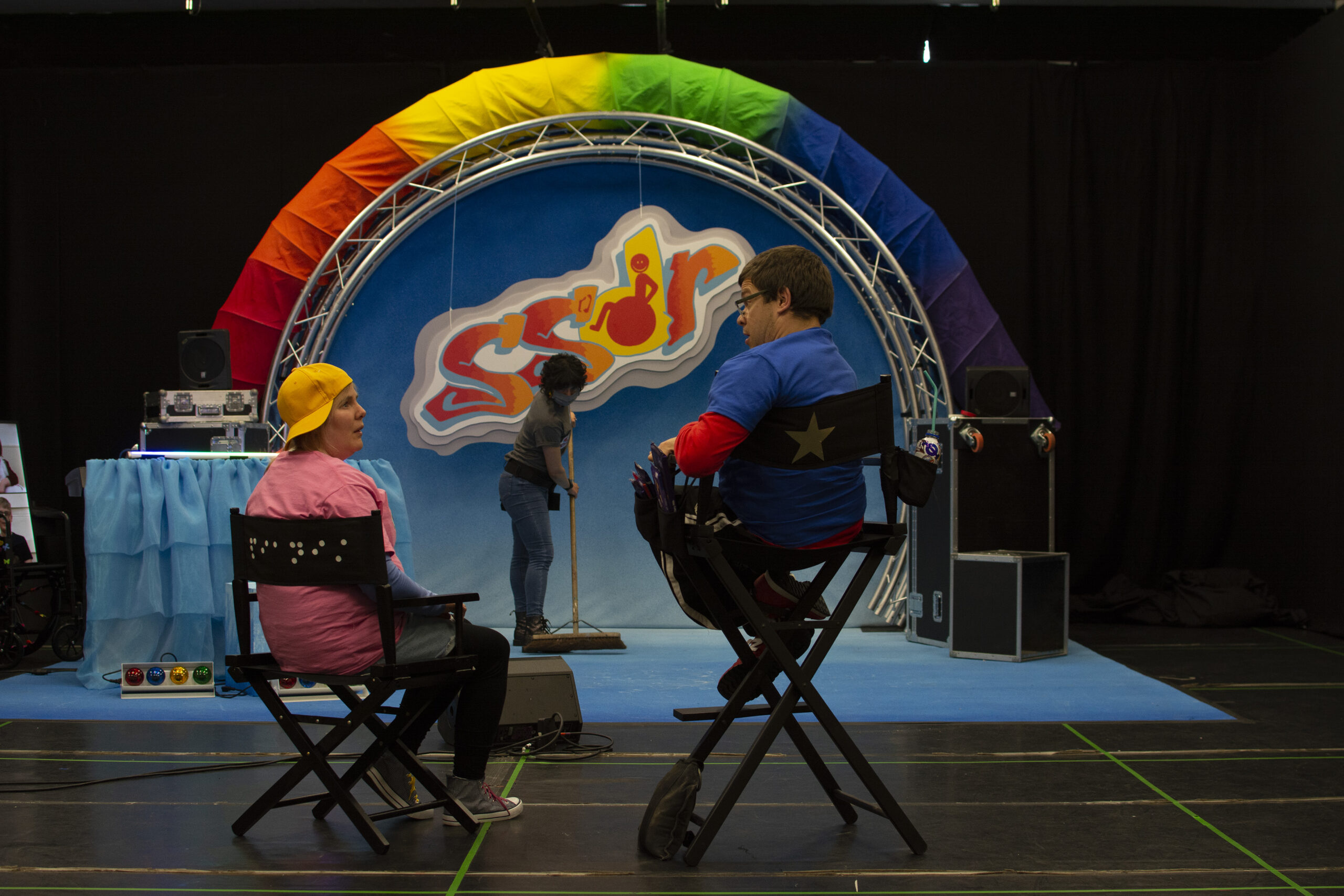 Robert and Sally are sitting in foldout chairs in front of the aforementioned set. His seat is much taller than hers. He is looking down at her as they appear to be having a conversation. On set there is a woman with short dark hair sweeping the stage.
