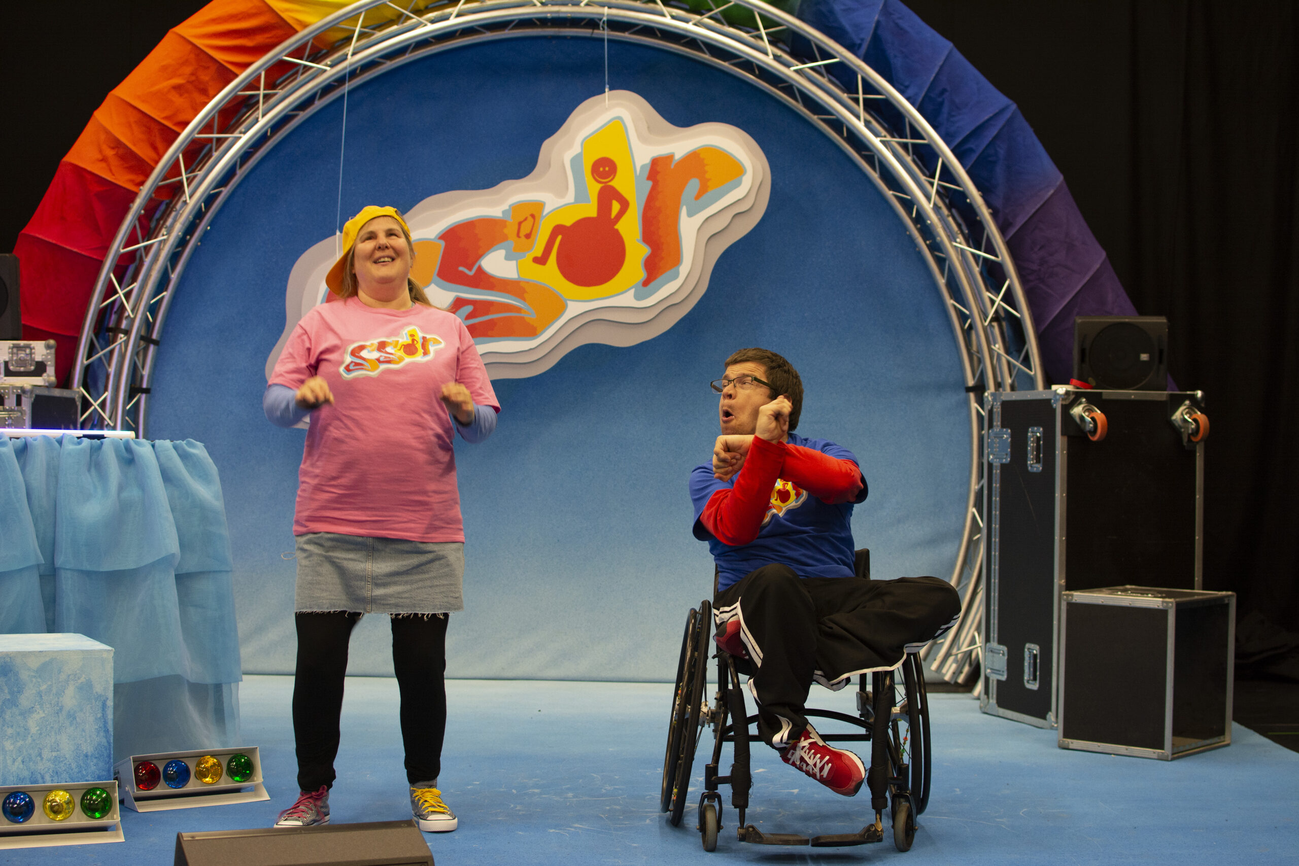 Sally and Robert are positioned at the front of the stage. Robert, on the right, has his arms positioned in an X as he looks over at Sally. Sally, on the left, is smiling and has her hands lifted at her sides as if mid gesture.