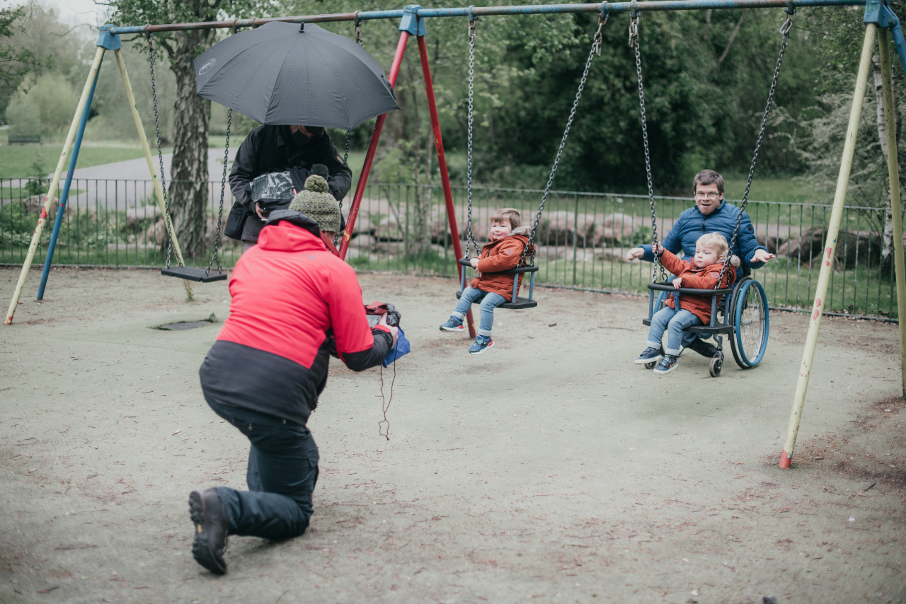 A swing set outside. There are two very young toddlers, each in their own swing with behind pushing them. In front of them there is a man on one knee holding a camera filming them. Everyone is wearing thick winter coats.