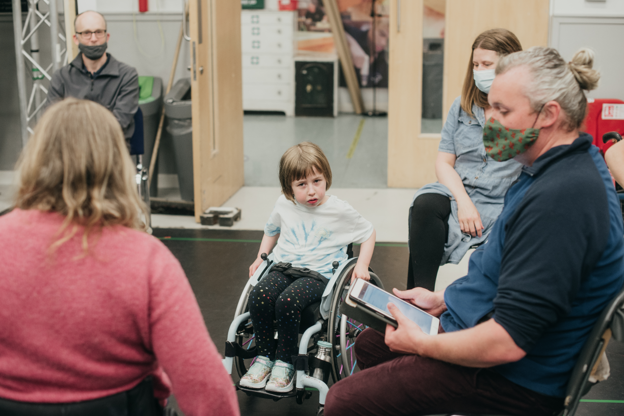 People sit around a rehearsal room. In the foreground is Oona in her chair, in the background is Joe, holding an iPad and Sal facing Oona with her back to the camera.