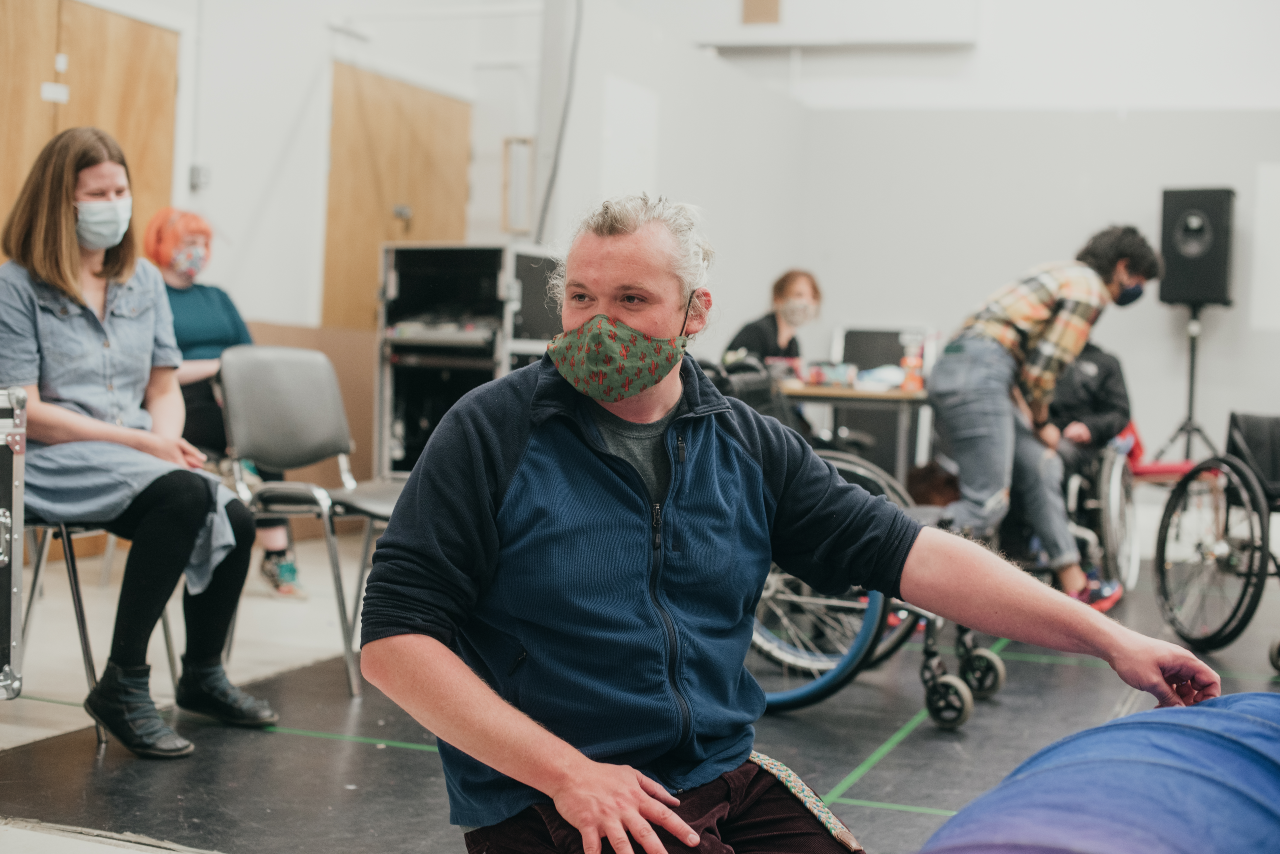A rehearsal room full of people spread out on chairs. The camera is focused on Joe (the director) middle aged man with long grey hair that is pulled back. He is wearing a green patterned mask and is looking at something off camera. He has one hand reached out and is touching something large and blue on the floor. 
