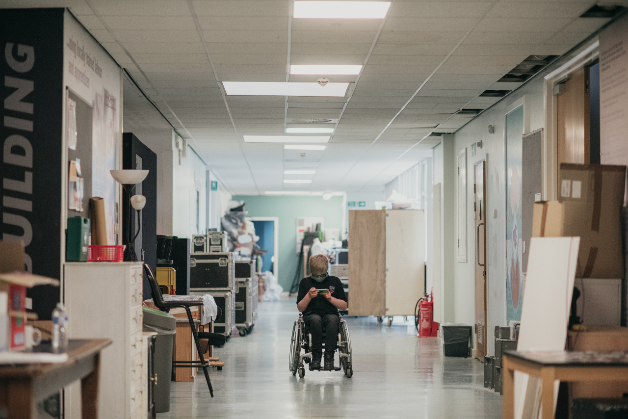 A shot of a cluttered corridor. There are various things lining it, such as: lights, cardboard boxes and pieces of furniture. In the middle there is Ollie in a wheelchair looking down at their phone. 