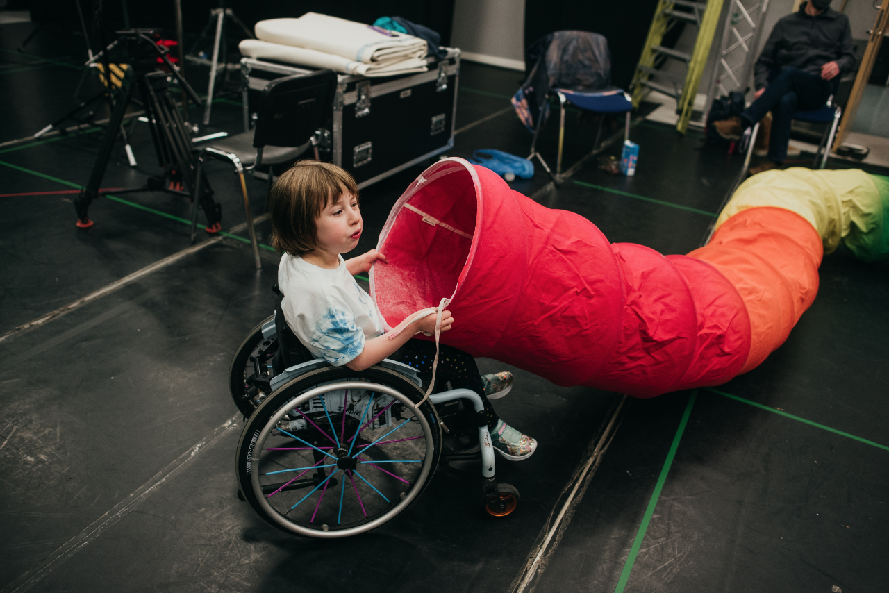Oona sits backstage. She is holding a large red and green tube in their hands as if about to dunk her head in the end. It is about large enough for a person to crawl through.