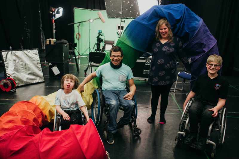 Sal, Rob, Oona and Ollie pose with big smiles, and smile for a photograph. They are backstage and there is lighting and sound equipment in the background all set against a greenscreen. They are posing with a large rainbow tube around them.
