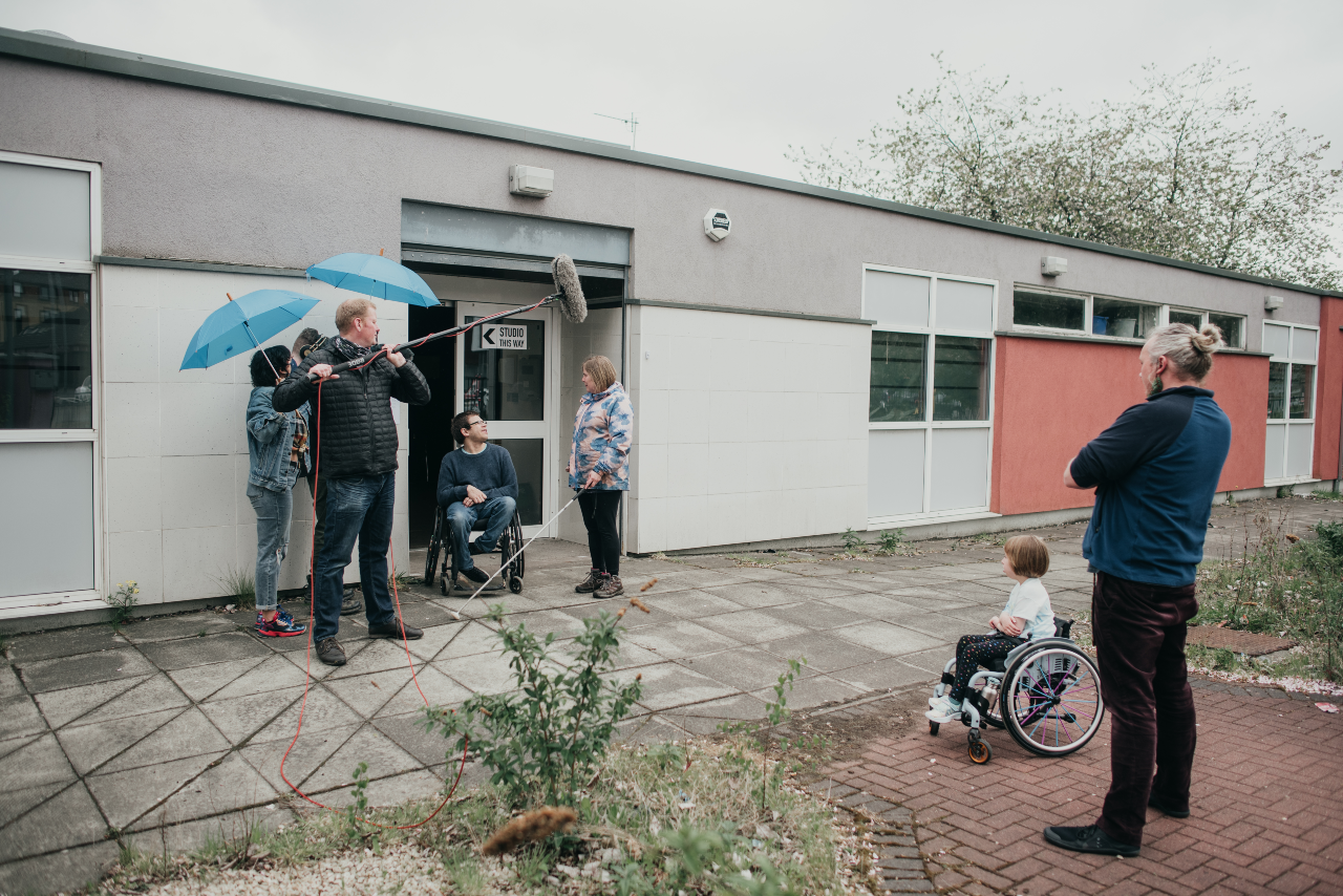 A photo taken outside of a small building. Rob and Sal are surrounded by a camera crew. There is a camera and a mic on a boom pointed at them. Joe and Oona watch on from a distance.