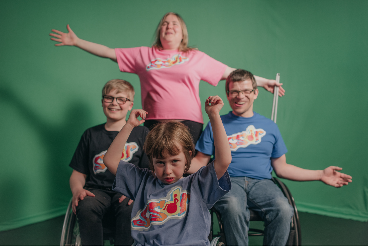 Sal, Rob, Ollie and Oona pose in front of a green screen in a diamond formation. They are all wearing different coloured T shirts that have ‘SSDR’ written on them. Sal is at the back, smiling with her arms outstretched and a folded cane in one hand. In front of her there are Roba dn Ollie, who are both smiling. At the very front is is Oona, who has her arms in the air.