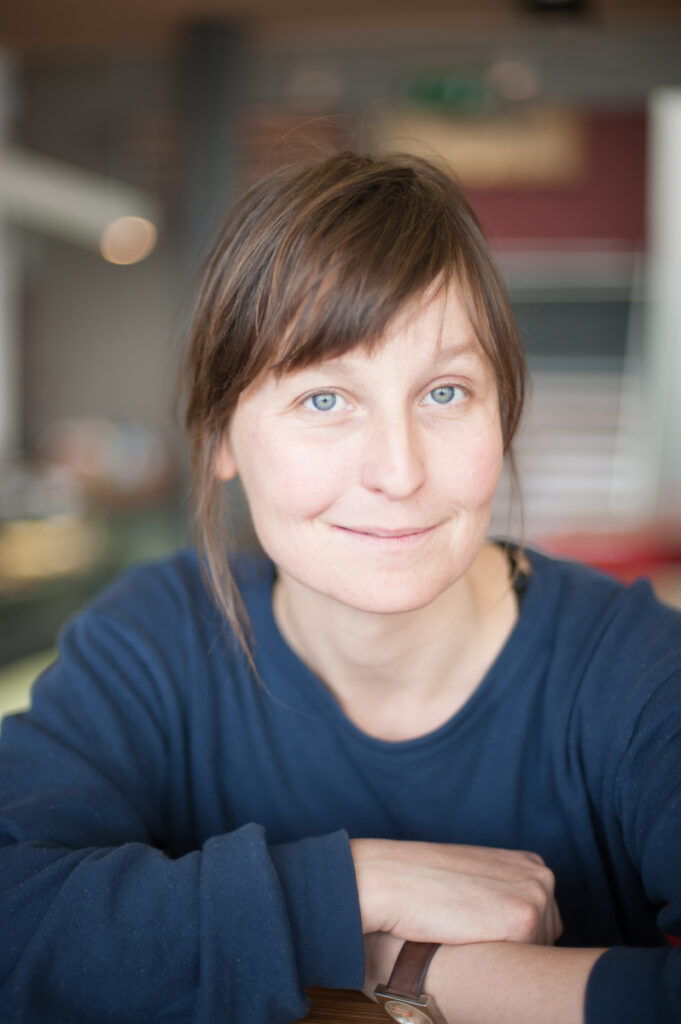 Lu is a white woman in her 50s. Her longish brown hair is tied back, with a few strands dangling past her ears. She sits, leaning her folded arms on a table, smiling inquisitively at the camera.