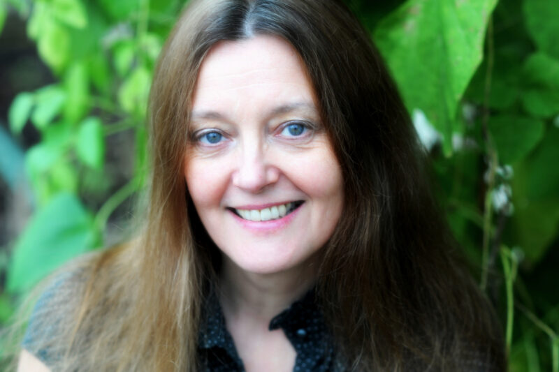 Gerda is a white woman in her 50s. Her brown hair is flecked with bits of grey and hangs down past her shoulders. She smiles widely into the camera, standing in front of a leafy background.