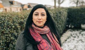 Niloo is a young woman of arabic heritage. She has long black hair and a nose ring, and looks into the camera in a contemplating manner. She wears a grey jumper and a red patterned scarf. She stands outdoors in a snowy garden, in front of a high hedge.