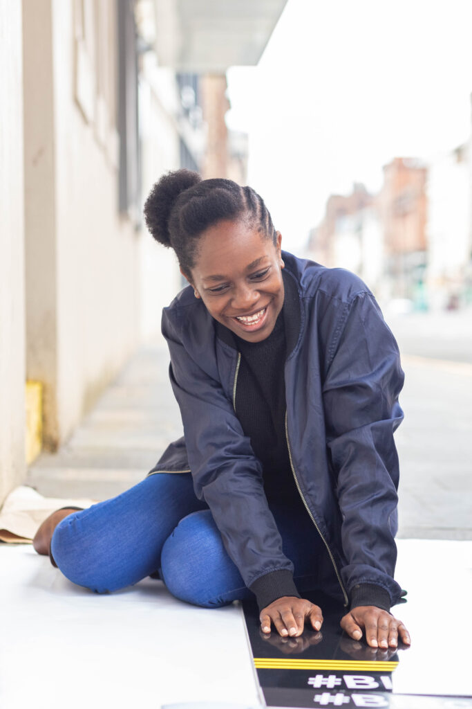 Wezi is a black woman in her 30s. She sits on the floor, wearing jeans and a bomber jacker. She is leaning over a Black Lives Matter placard and smiling widely.