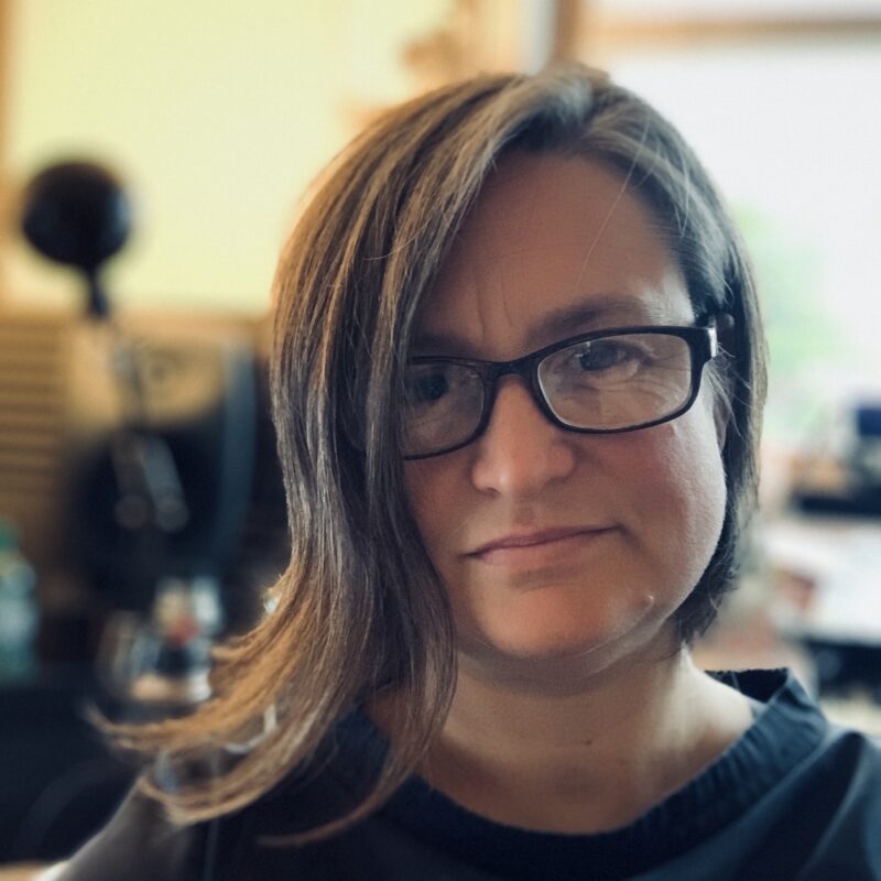 Zoe Irvine is a white woman in her 50s. She wears glasses and her medium length hair is swept over the right side of her face in an arc that touchers her chin.