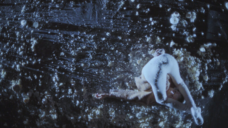 Photo from birds eye perspective. The black floor is scattered with white dusty lumps varying in size from very small to about the size of a tennis ball. A low side light catches the detritus on the floor, casting long shadows across the plane. It looks like the surface of the moon. A figure off to the right of the photo, is dressed in neutral white leggings and a light brown cardigan. They have rocked up onto their shoulders, their bum in the air and their knees framing their face, which stares neutrally up into space. Their left arm stretches out into the dappled floor.