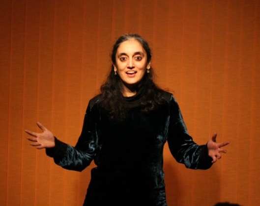 Gauri is a woman of Asian heritage, in this photo in her early 30s. She stands on a stage with her arms spread out wide and her fingers open. She is telling a story to an audience. She stands in front of an organe curtain. She has long dark hair that falls past her shoulders and small earrings in.