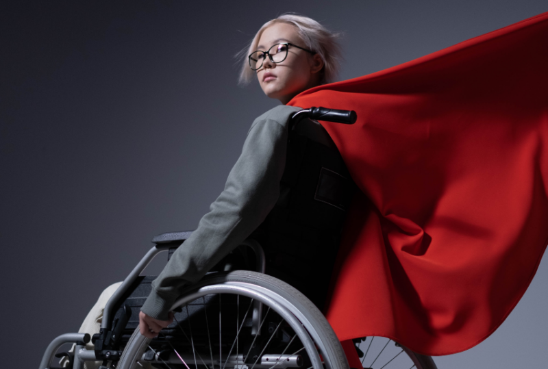 A young Asian woman sits in a wheelchair, facing away from the camera, but with her head turned over her left shoulder to face the camera. A red cape billows off from her neck line, rippling in the wind.
