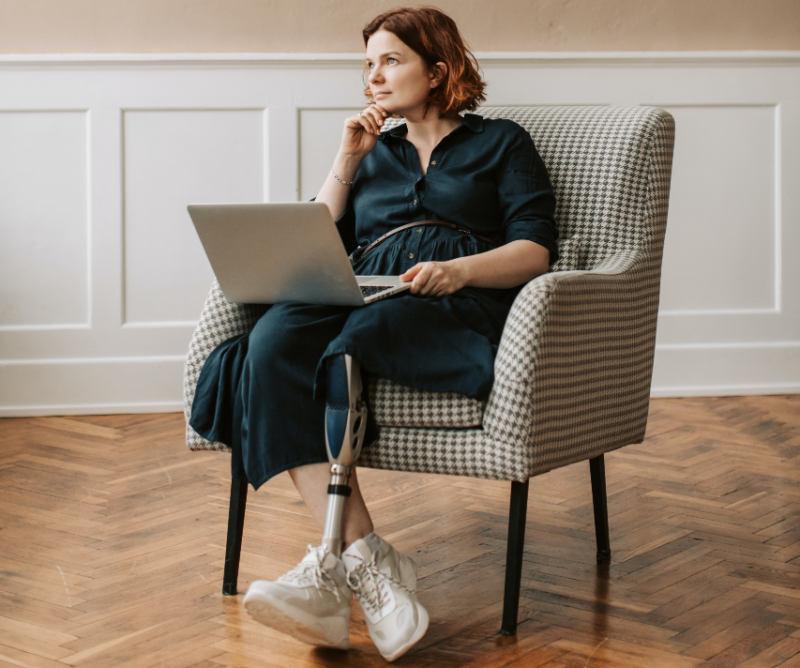 A young woman sits on a comfy chair looking thoughtfully into the distance. Her right hands props up her chin. Her legs are corssed and her left leg is a prosthetic leg. Balanced on her lap is a laptop.