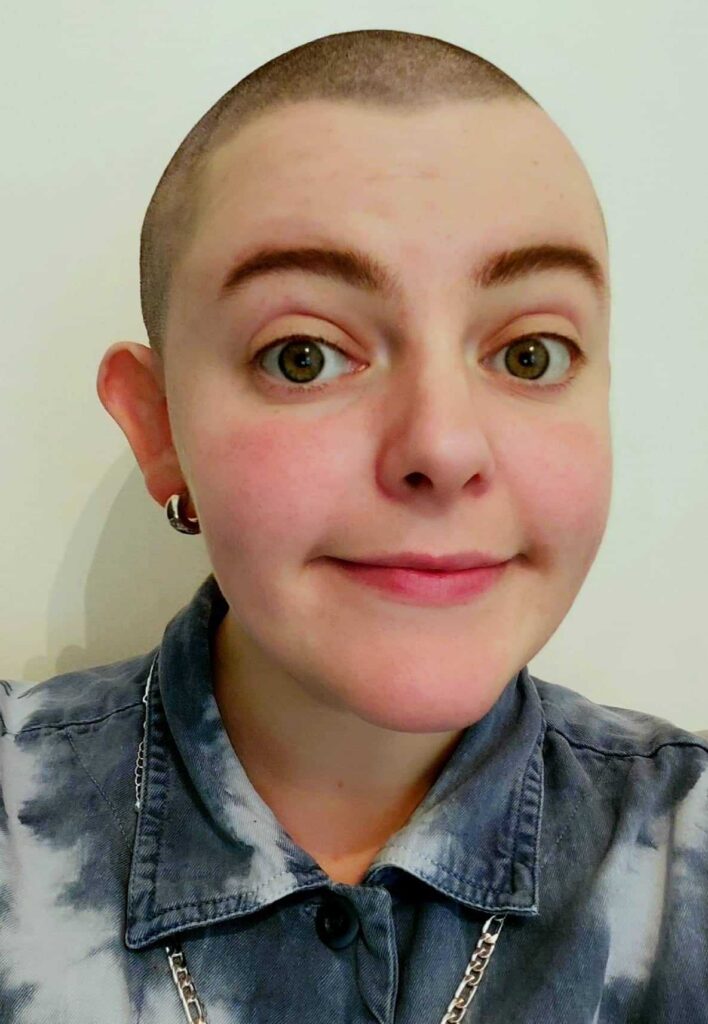 Headshot of Nelly Kelly. A white person in their late 20s. Wearing a blue denim shirt and with a close shaved head buzzcut. With a open and welcoming face.