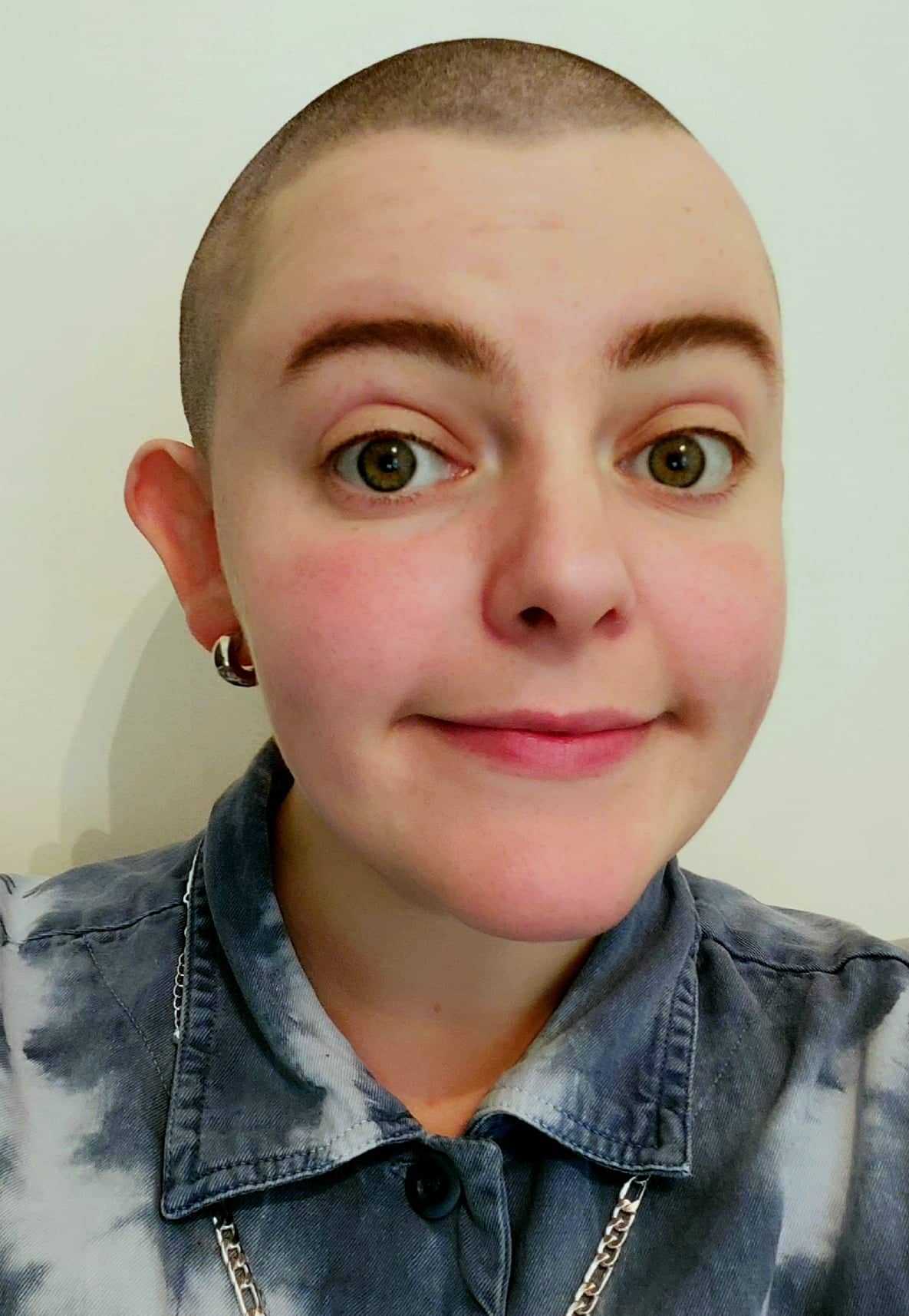 Headshot of Nelly Kelly. A white person in their late 20s. Wearing a blue denim shirt and with a close shaved head buzzcut. With a open and welcoming face.