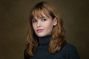 Headshot of Hannah Yayha Hassan: An arabic woman in her early 30s. She is wearing a black polo neck and has shoulder length golden brown hair and gives a gentle smile to camera.